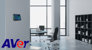 A sleek office space featuring a mounted AVer device on the wall, a round table with a laptop, and black ergonomic chairs, highlighted by large windows showcasing a cityscape.